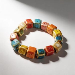 Op de afbeelding: Een armband bestaande uit kubusvormige kralen in verschillende kleuren. De kralen zijn geglazuurd in tinten rood, oranje, geel, blauw en crème. De armband is een uniek sieraad.