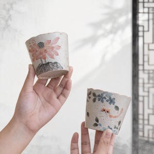 Könnte beinhalten: Zwei Keramiktöpfe mit gewellten Rändern werden hochgehalten. Einer der Töpfe zeigt ein rosa Lotusblumen-Design, der andere ein Katzen- und Blumenmuster in Blau und Orange. Die Töpfe haben einen neutralen, gesprenkelten Hintergrund.