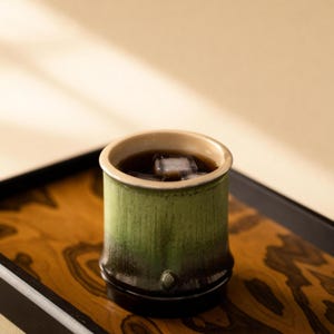 May include: A green bamboo-style cup containing a dark liquid and ice cubes, presented on a wooden tray with a black border. The cup features a light beige rim and a dark base. The tray has a wood grain pattern.
