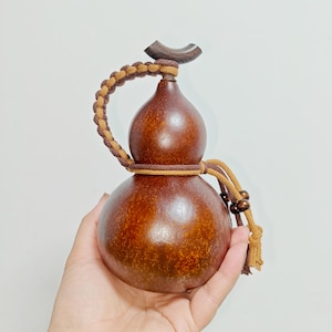 Peut inclure: Un récipient en forme de gourde marron avec une finition lisse et polie. Il est surmonté d'un bouchon en bois foncé et orné d'un cordon tressé beige et marron foncé qui entoure le goulot et se termine par de petites perles en bois.