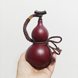 Peut inclure: Un récipient en forme de gourde rouge bordeaux profond avec une finition lisse. Il présente une anse en corde tressée marron foncé, un petit accent carré doré avec un motif géométrique et un bouchon noir incurvé au sommet, noué d'un cordon marron.