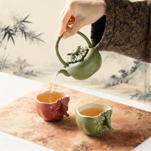 以下が含まれることがあります： 葉の形をした蓋が付いた緑色のセラミックティーポットが、2つの小さな装飾カップに紅茶を注いでいます。カップの1つは赤、もう1つは緑です。カップとティーポットは模様のある表面にあり、背景は絵画です。