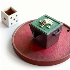 Könnte beinhalten: Eine würfelförmige Tasse mit Flüssigkeit steht neben einem Miniatur-Spielset auf einem roten Tablett. Das Spielset ist eine braune Box mit grünem Deckel, Würfeln und kleinen Spielsteinen. Die Tasse ist weiß mit schwarzen und orangefarbenen Punkten.