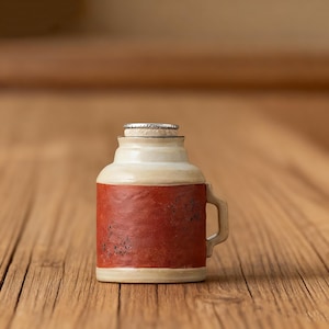 Op de afbeelding: Een keramische flesopener met een handvat, met een crèmekleurige bovenkant en een donkerrood rechthoekig etiket. Een flesdop zit erop. De opener staat op een houten oppervlak.