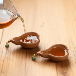Könnte beinhalten: Zwei kleine, braune, birnenförmige Teetassen mit einer dekorativen Perle am Griff. Tee wird aus einer Glaskanne in eine der Tassen gegossen. Die Tassen sind mit Tee gefüllt.