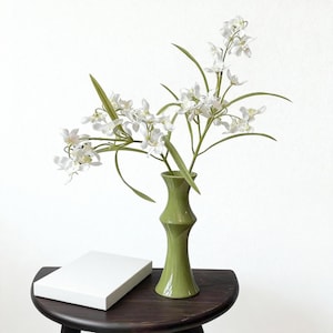May include: A green bamboo-shaped vase holds white orchid stems with green leaves. The vase sits on a dark wooden table next to a white rectangular box. The background is a plain white wall.
