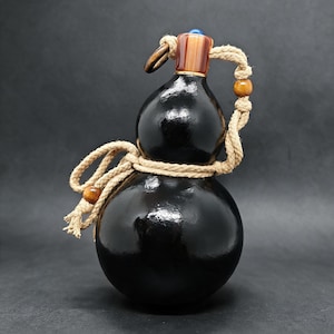 Könnte beinhalten: Eine schwarze, kürbisförmige Flasche mit glänzender Oberfläche und einem beigefarbenen Seilgriff, der um den Hals und Körper gewickelt ist. Der Verschluss besteht aus braunem und bernsteinfarbenem Material mit einer kleinen blauen Perle oben.