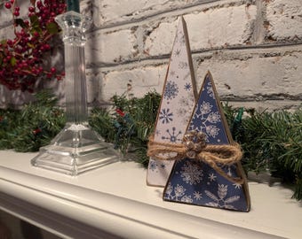 May include: Two triangular Christmas tree decorations. One is white with blue snowflakes, the other is blue with white snowflakes. Both are wrapped with twine and a decorative silver embellishment. The trees are on a white surface with a brick background.