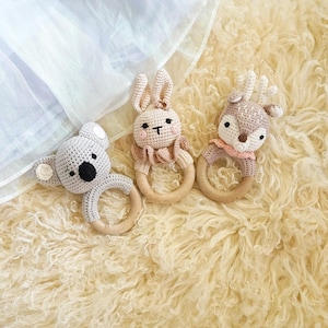 May include: Three crochet baby rattles in the shape of a koala, bunny, and deer. Each rattle has a wooden ring and is made of soft, neutral-colored yarn. The koala is gray, the bunny is beige, and the deer is light brown.