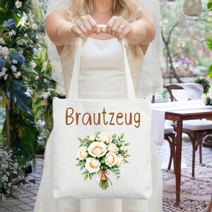 Könnte beinhalten: Eine weiße Tragetasche mit dem Wort "Brautzeug" in braunen Buchstaben und einer floralen Bouquet-Illustration. Das Bouquet zeigt weiße Rosen und grüne Blätter. Die Tasche wird von einer Person gehalten, die ein Hochzeitskleid und einen Schleier trägt.