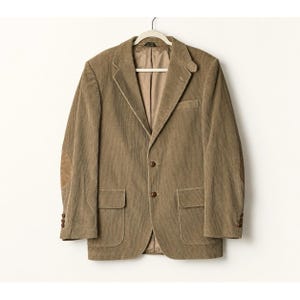 May include: A tan corduroy blazer featuring a notched lapel, two front buttons, two flap pockets, and suede elbow patches. The jacket has long sleeves with three buttons at each cuff and a smooth, light brown interior lining visible at the collar.