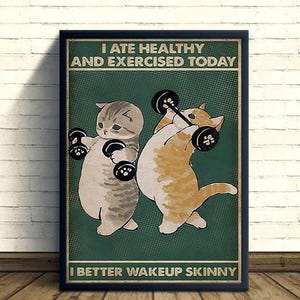 Könnte beinhalten: Ein gerahmtes Poster mit dunkelgrünem Hintergrund zeigt zwei Cartoon-Katzen, die schwarze Hanteln heben. Der Text oben lautet I ATE HEALTHY AND EXERCISED TODAY und der Text unten I BETTER WAKEUP SKINNY. Die Katzen sind grau und orange gefärbt.