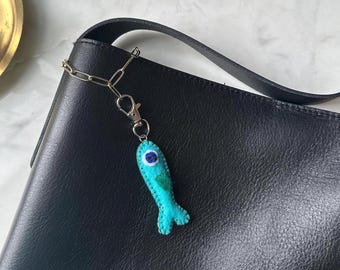 May include: A teal felt fish keychain with a blue and white eye, attached to a silver chain and clasp. The keychain hangs from the black strap of a black leather bag. The fish has a small green accent.