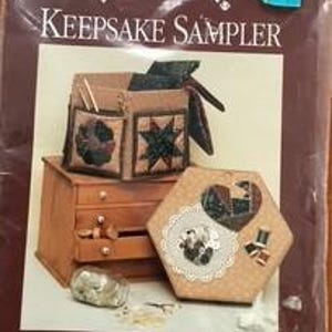 May include: A Keepsake Sampler kit by Dream Spinners. The image shows a wooden chest, a hexagonal box with a heart design, and a brown fabric bag with pockets. The kit includes patterns and directions for the items.