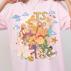 Op de afbeelding: Een lichtroze t-shirt met een grafische print van meerdere geanimeerde personages in diverse outfits, waaronder ruimte- en westernstijlen. Het ontwerp bevat blauwe wolken, een raket, een sheriffster en de grote gele letters TS.