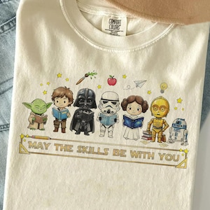 以下が含まれることがあります： クリーム色のTシャツ。スターウォーズのキャラクターが本を読んでいるグラフィックがプリントされています。「MAY THE SKILLS BE WITH YOU」の文字が下に印刷されています。Comfort Colors製です。