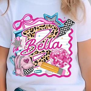 Pode incluir: Uma t-shirt branca de manga curta com um design gráfico que apresenta uma letra B grande com estampado de leopardo, o nome Bella em letra cursiva rosa, uma mochila rosa, um lápis amarelo, uma bola de discoteca rosa, um laço rosa e xadrez.