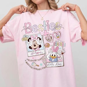 Peut inclure: T-shirt rose clair avec le mot "Besties" en couleurs pastel. Le t-shirt pr&eacute;sente Minnie Mouse et Daisy Duck dans des cadres de style polaroid, avec des c&oelig;urs, des biscuits et de la cr&egrave;me glac&eacute;e. Le t-shirt a une coupe ample.