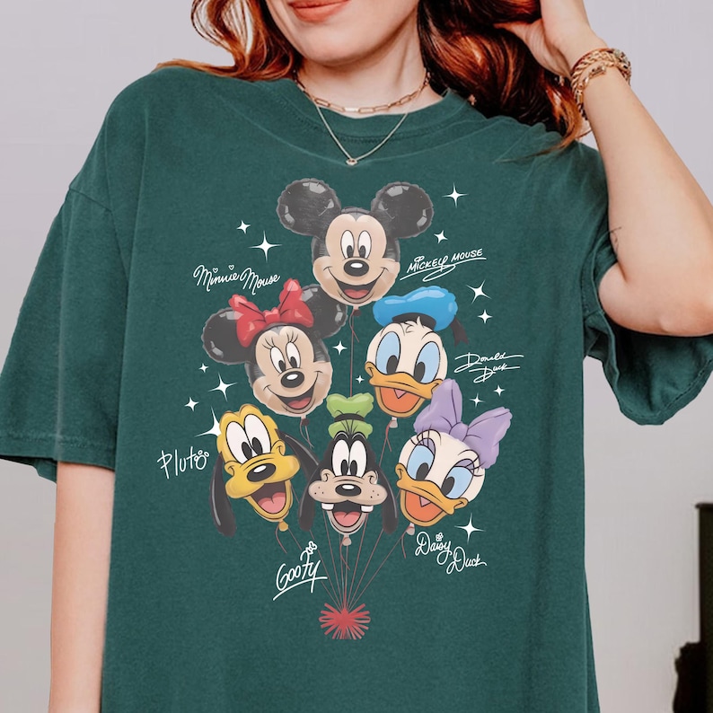 May include: Dark green t-shirt featuring a colorful graphic of Disney characters as balloons. Mickey Mouse, Minnie Mouse, Donald Duck, Daisy Duck, Goofy, and Pluto are depicted. The names of the characters are written in a script font.