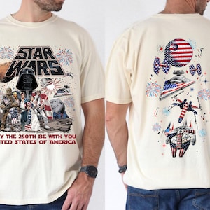 以下が含まれることがあります： アメリカ国旗柄のスター・ウォーズのキャラクターや乗り物が描かれたクリーム色のTシャツ。前面には「MAY THE 250TH BE WITH YOU UNITED STATES OF AMERICA」という文字と、ダース・ベイダー、ルーク・スカイウォーカー、チューバッカがプリントされています。