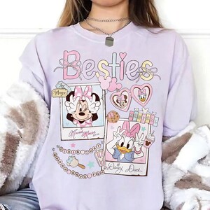 Peut inclure: Un sweat-shirt lilas clair avec le mot "Besties" dans une police fantaisie. Le motif comprend des images de Minnie Mouse et Daisy Duck, avec les mots "Magic" et "Magical". Le sweat-shirt a une coupe ample.