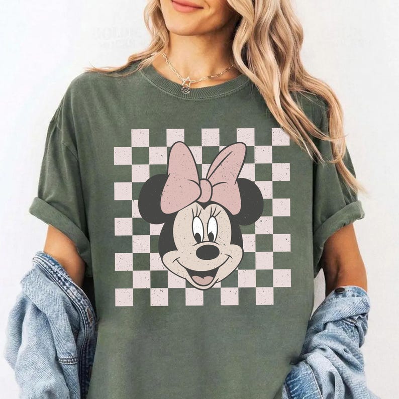 May include: Olive green t-shirt featuring a Minnie Mouse graphic with a pink bow and a checkered background. The t-shirt has rolled-up sleeves and a relaxed fit. The graphic is centered on the front of the shirt.