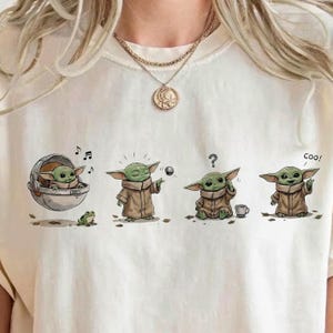 Op de afbeelding: Crèmekleurig T-shirt met een cartoonafbeelding van een groen, buitenaards personage in verschillende poses. Het personage heeft grote oren en een bruine mantel. Het shirt heeft een ronde hals en korte mouwen.