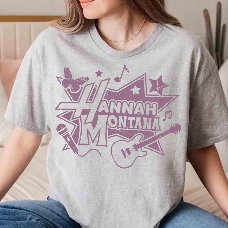 May include: Gray t-shirt featuring a purple graphic design with the text "Hannah Montana", a butterfly, musical notes, a microphone, a star, and a guitar. The shirt is a light gray color.
