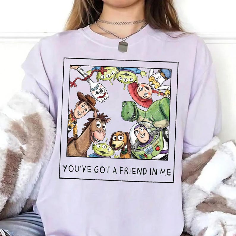 May include: A light purple sweatshirt featuring a framed graphic of characters from the animated film "Toy Story." The graphic includes Woody, Buzz Lightyear, Rex, and the Little Green Men. The text "YOU'VE GOT A FRIEND IN ME" is printed below the graphic.