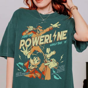 May include: Teal green t-shirt featuring a graphic of cartoon characters with the text "POWERLINE WORLD TOUR '95". The design includes the text "STAND OUT" and song lyrics. The shirt has a vintage, distressed look.