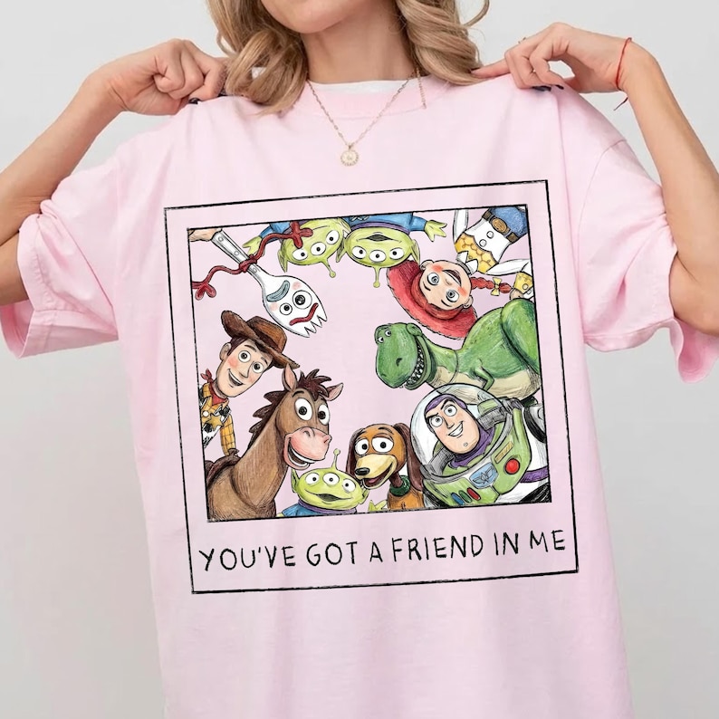 May include: A light pink t-shirt featuring a framed graphic of Toy Story characters. The graphic includes Woody, Buzz Lightyear, and other characters. The text "YOU'VE GOT A FRIEND IN ME" is printed below the graphic.