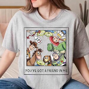 May include: A light gray t-shirt featuring a graphic of characters from the animated film Toy Story. The graphic includes Woody, Buzz Lightyear, Rex, and the Little Green Men, with the text "YOU'VE GOT A FRIEND IN ME" in a black rectangle.