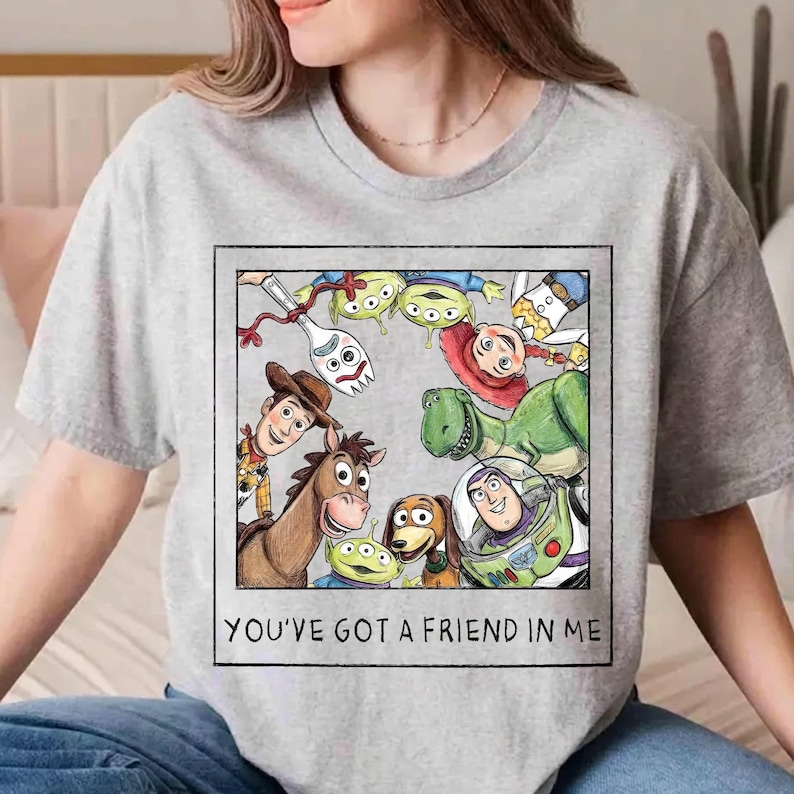 May include: A light gray t-shirt featuring a graphic of characters from the animated film Toy Story. The graphic includes Woody, Buzz Lightyear, Rex, and the Little Green Men, with the text "YOU'VE GOT A FRIEND IN ME" in a black rectangle.