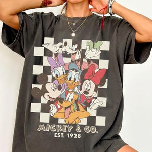 May include: Dark gray t-shirt featuring a graphic of Mickey Mouse and friends on a checkered background. The design includes Mickey, Minnie, Donald, Daisy, Goofy, and Pluto. The text "Mickey & Co. Est. 1928" is printed below the characters.