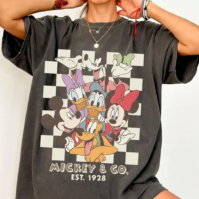May include: Dark gray t-shirt featuring a graphic of Mickey Mouse and friends on a checkered background. The design includes Mickey, Minnie, Donald, Daisy, Goofy, and Pluto. The text "Mickey & Co. Est. 1928" is printed below the characters.