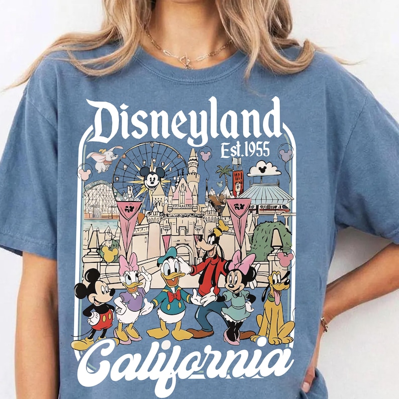 May include: Blue t-shirt featuring a Disneyland California graphic. The design includes Mickey Mouse, Minnie Mouse, Donald Duck, Goofy, and Daisy Duck, along with iconic park imagery. The text "Disneyland Est.1955" is displayed above the park illustration.