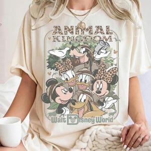 May include: Cream-colored t-shirt featuring a graphic of Disney characters, including Mickey, Minnie, Donald, Daisy, Goofy, and Pluto, in safari attire. The text "ANIMAL KINGDOM" and "Walt Disney World" are printed on the shirt.