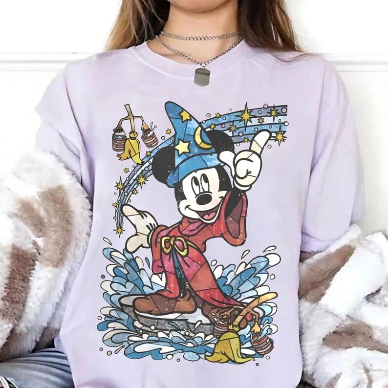 Pu&ograve; includere: Una t-shirt viola chiaro con una grafica di Topolino vestito da mago con una tunica rossa e un cappello blu con stelle gialle e una mezzaluna. Il design include una scopa gialla, secchi d'acqua e spruzzi d'acqua di colore blu.