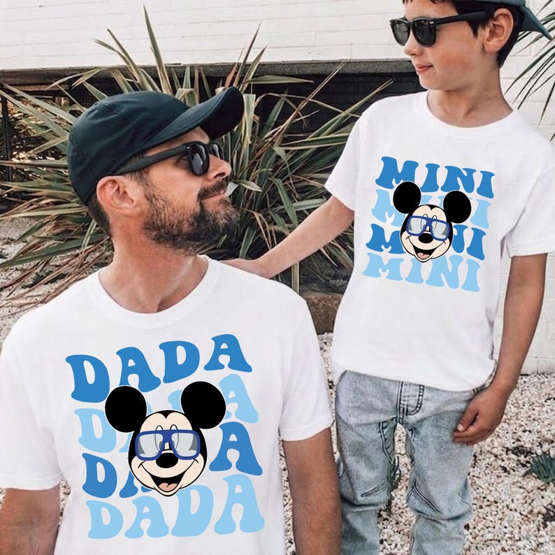 K&ouml;nnte beinhalten: Zwei wei&szlig;e T-Shirts mit blauem Text und einer Mickey Mouse-Grafik. Eines sagt "DADA" und das andere "MINI". Beide Shirts zeigen die Zeichentrickfigur mit Brille. Die Shirts werden von zwei Personen getragen.