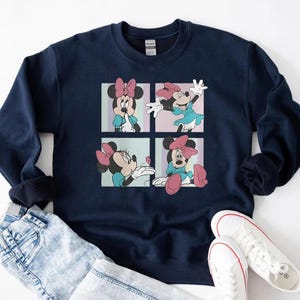 May include: Navy blue sweatshirt featuring a four-panel graphic of Minnie Mouse in various poses. Minnie Mouse is wearing a pink bow and a blue dress in each panel. The sweatshirt is paired with light blue jeans and white sneakers.