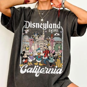 May include: Dark gray t-shirt featuring a colorful Disneyland graphic. The graphic includes Mickey Mouse, Donald Duck, Goofy, and other Disney characters, along with the words "Disneyland California Est. 1955."