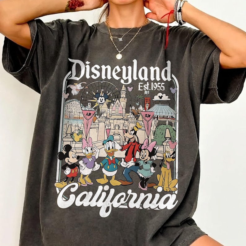 May include: Dark gray t-shirt featuring a colorful Disneyland graphic. The graphic includes Mickey Mouse, Donald Duck, Goofy, and other Disney characters, along with the words "Disneyland California Est. 1955."