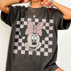 May include: Dark gray t-shirt featuring a Minnie Mouse graphic with a pink bow and a checkered background. The graphic tee has a vintage, distressed look.