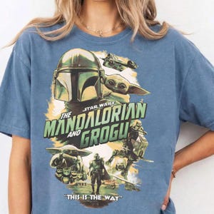 May include: Blue t-shirt featuring a graphic design from Star Wars: The Mandalorian. The design includes the Mandalorian's helmet, Grogu, and the text "The Mandalorian and Grogu" and "This is the Way".