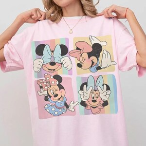 May include: A light pink t-shirt featuring four panels with different Minnie Mouse illustrations. Each panel has a pastel rainbow striped background. Minnie Mouse is wearing a bow in each panel.