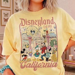 May include: Yellow t-shirt featuring a Disneyland California graphic. The design includes Mickey Mouse, Minnie Mouse, Donald Duck, Goofy, and Pluto, along with the Disneyland logo and the year 1955. The graphic is in a vintage style.
