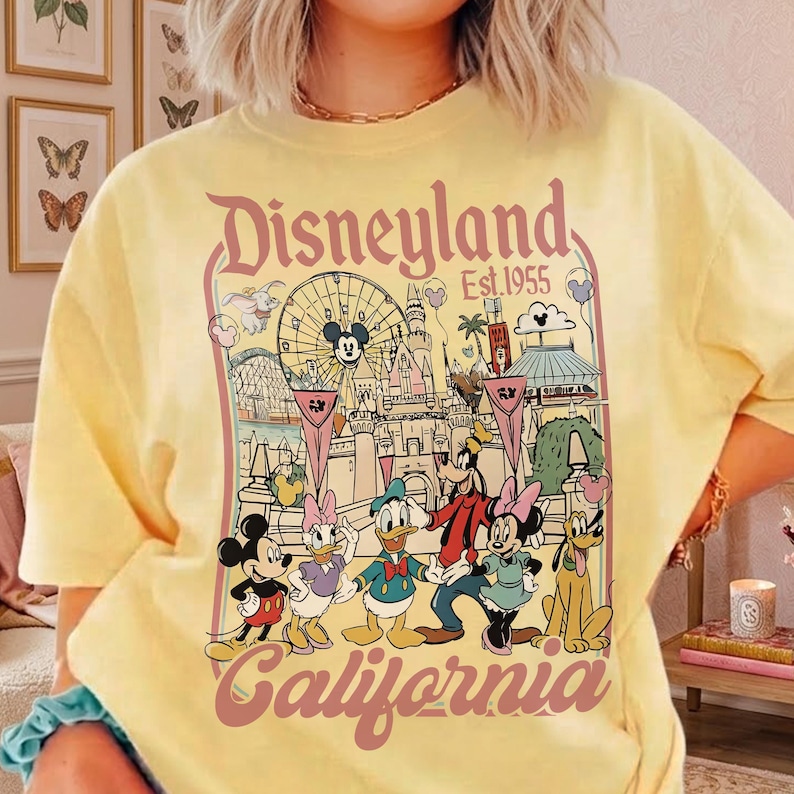 May include: Yellow t-shirt featuring a Disneyland California graphic. The design includes Mickey Mouse, Minnie Mouse, Donald Duck, Goofy, and Pluto, along with the Disneyland logo and the year 1955. The graphic is in a vintage style.