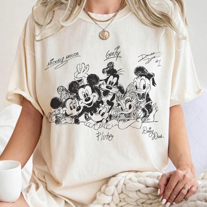Pu&ograve; includere: T-shirt color crema con un'illustrazione in bianco e nero di Topolino, Minnie, Pippo, Paperino, Paperina e Pluto. Le firme dei personaggi sono sopra o sotto. La maglietta ha maniche corte.