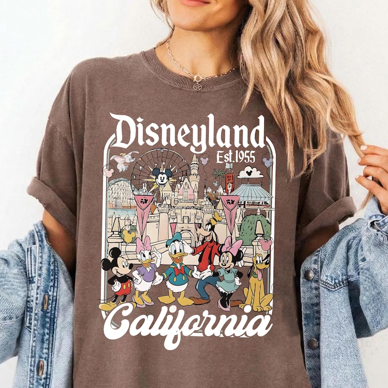 May include: Brown t-shirt featuring a Disneyland California graphic. The design includes Mickey Mouse, Minnie Mouse, Donald Duck, Goofy, and Daisy Duck, with the Disneyland castle and Ferris wheel in the background. The text "Disneyland Est. 1955" is also visible.