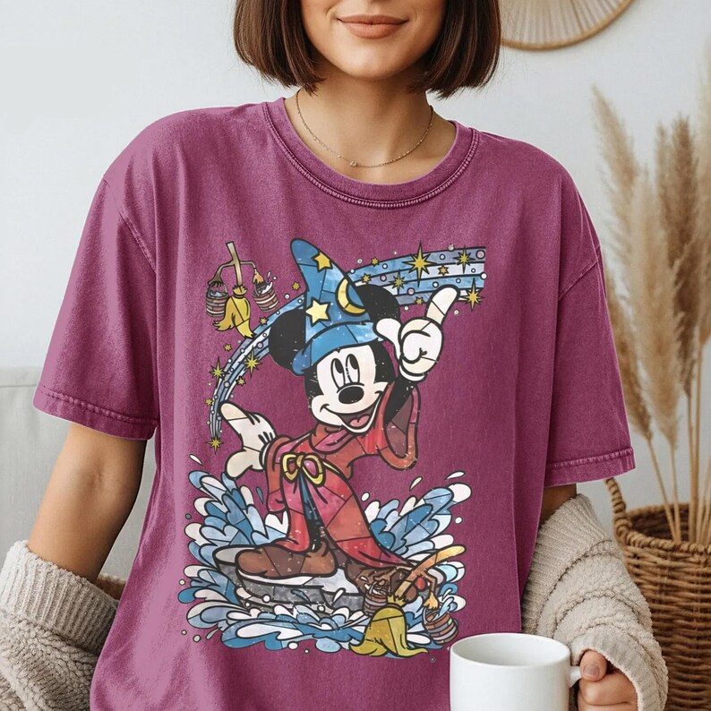 Pu&ograve; includere: Una t-shirt a maniche corte color magenta scuro con una stampa grafica di Topolino nelle vesti di stregone, con un cappello blu stellato e una tunica rossa, circondato da spruzzi d'acqua blu, scope gialle e una scia di stelle.